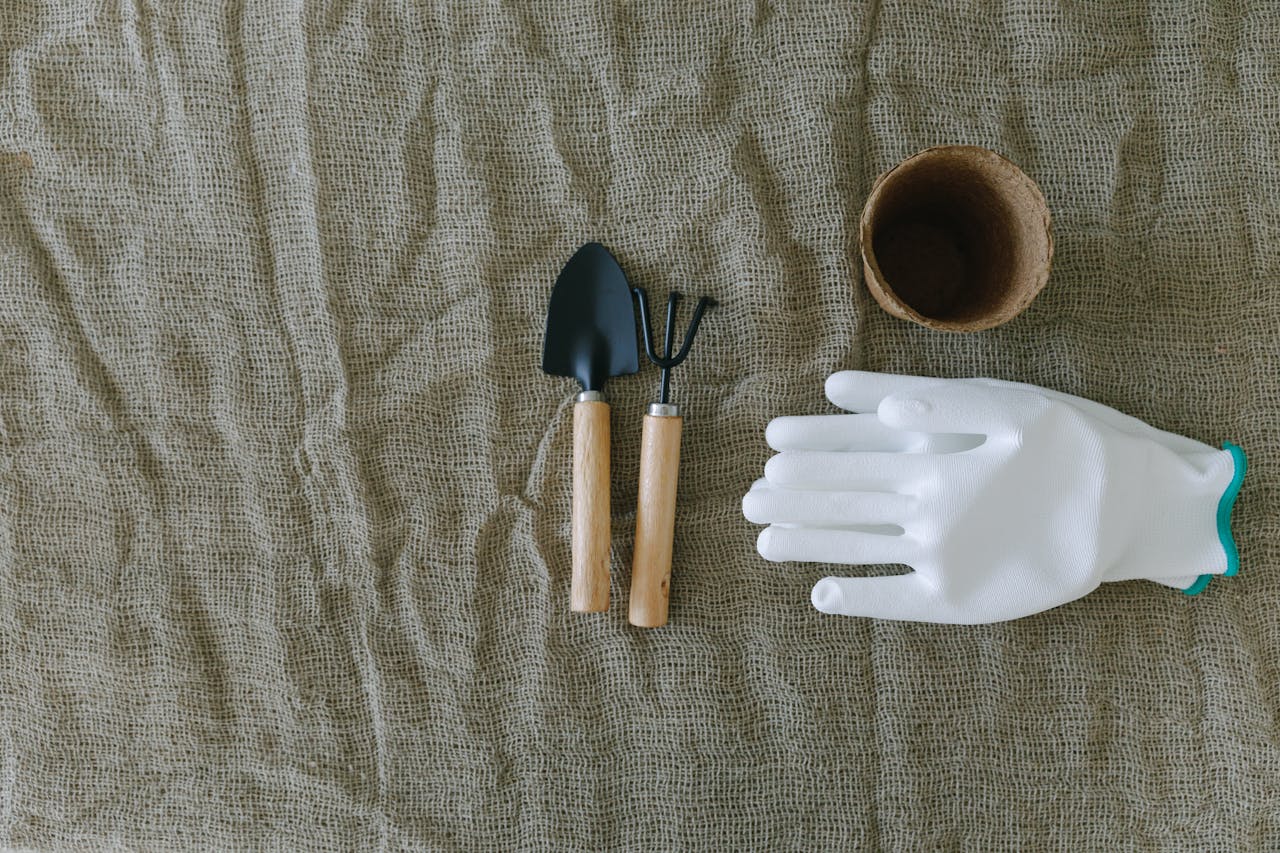 Flat lay of gardening tools, gloves, and pot on burlap surface.